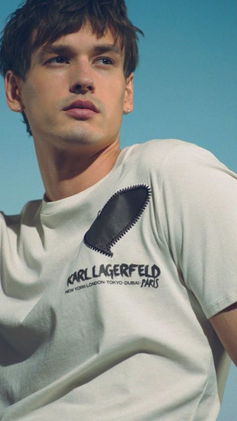 Close-up studio portrait of a young adult man wearing a light-colored Karl Lagerfeld t-shirt, against a plain blue backdrop, fashion shoot.