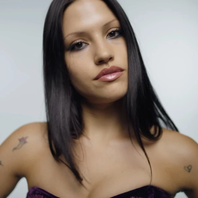 Close-up portrait of a woman with dark hair wearing a strapless purple top against a neutral backdrop, showcasing makeup and tattoos.