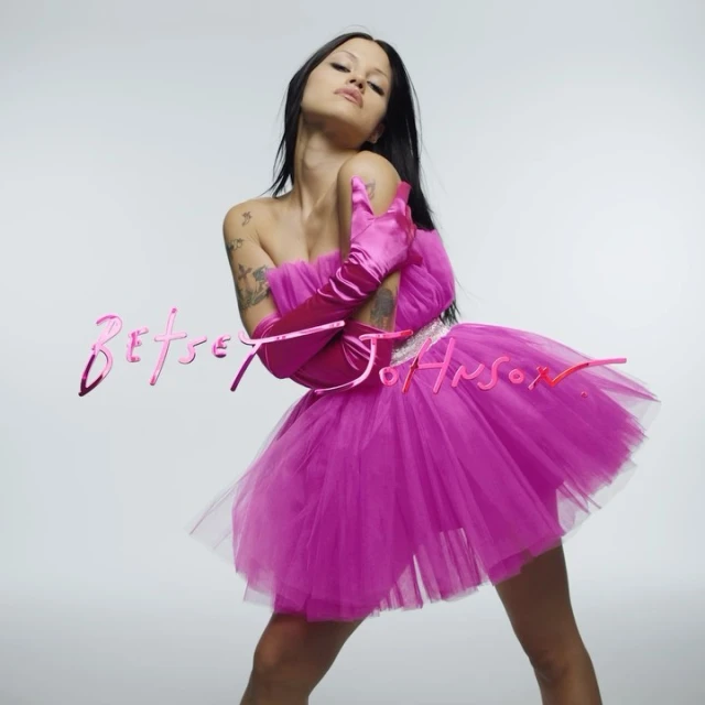 Woman in a pink tulle dress and gloves poses against a white background, featuring the Betsey Johnson logo. Fashion editorial image.