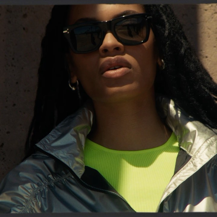 Close-up of a woman with dark hair and sunglasses, wearing a bright yellow top and iridescent jacket, standing in front of a wall.