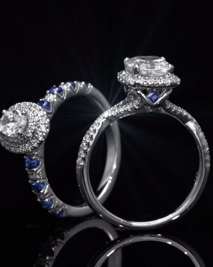 Close-up studio shot of two diamond engagement rings with white and blue gems on a glossy black surface and a dark background.