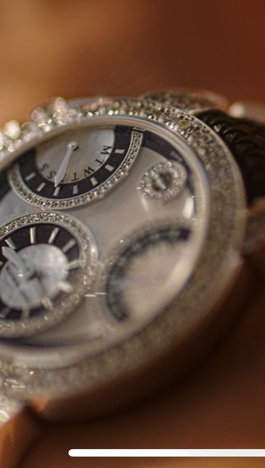 Close-up of an ornate wristwatch with diamonds. The watch face is light-colored with intricate detailing. The watch band is not visible.
