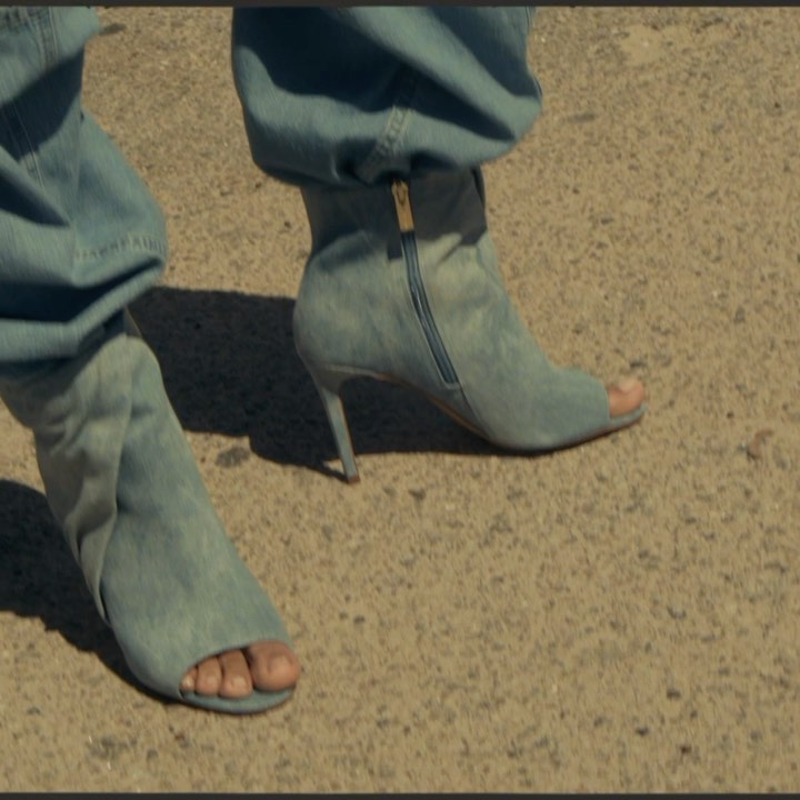 Fashionable peep-toe stiletto booties in light blue denim-like material, worn with matching baggy jeans, stand out against a grainy background.
