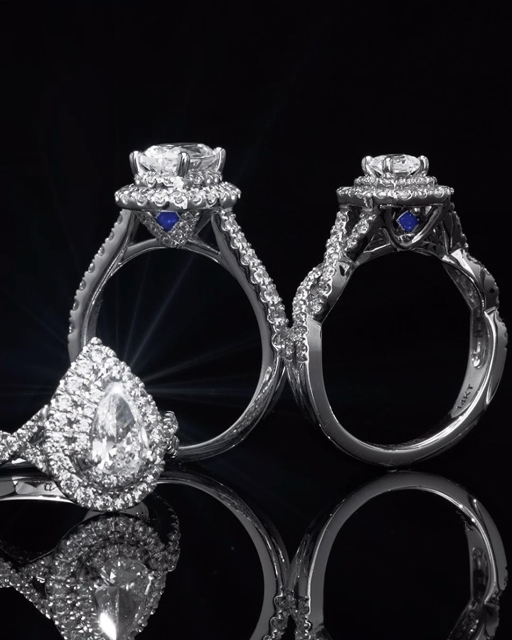 Close-up of three diamond engagement rings with platinum bands and accent stones against a black background with reflective surfaces.