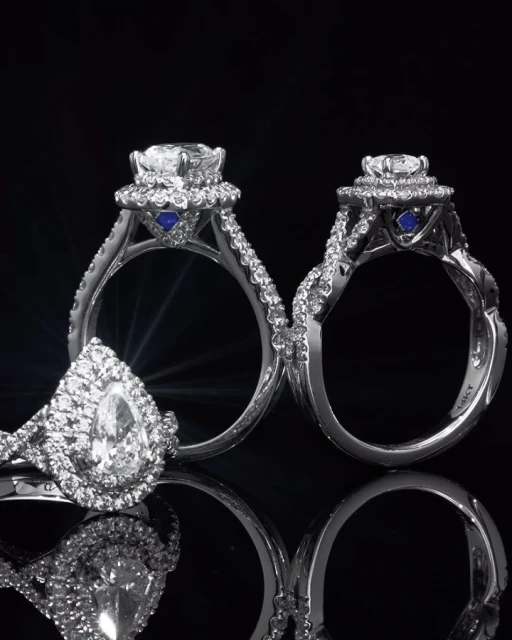 Close-up of three diamond engagement rings with platinum bands and accent stones against a black background with reflective surfaces.