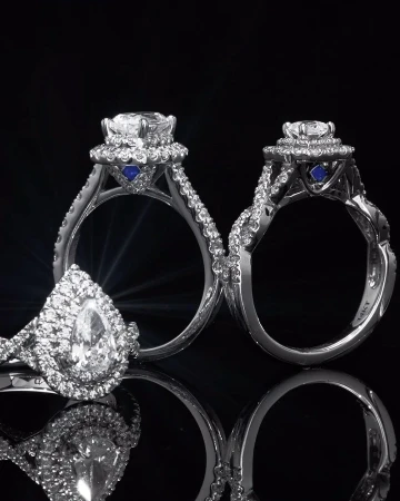 Close-up of three diamond engagement rings with platinum bands and accent stones against a black background with reflective surfaces.
