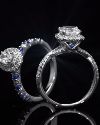Close-up product photography of two diamond and sapphire engagement rings on a dark reflective surface by Theo Darling