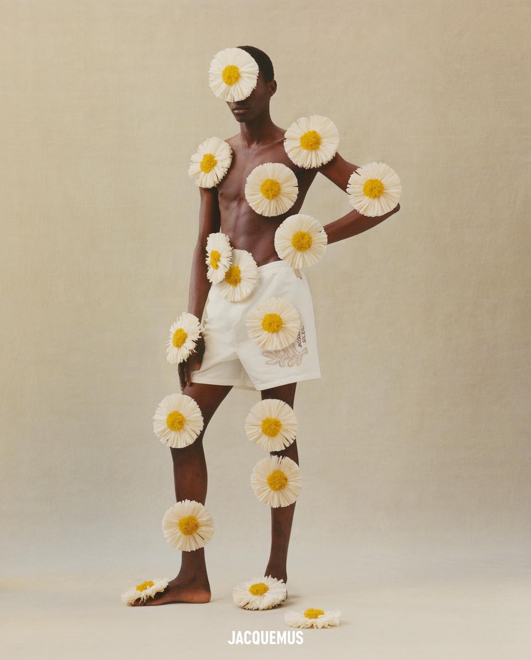 A Black male fashion model poses with daisies on a beige background, wearing white shorts. Conceptual studio photography.