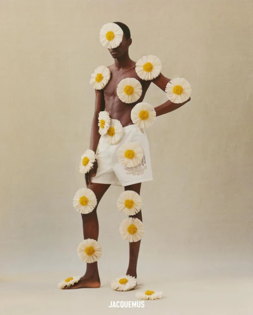 A Black male fashion model poses with daisies on a beige background, wearing white shorts. Conceptual studio photography.