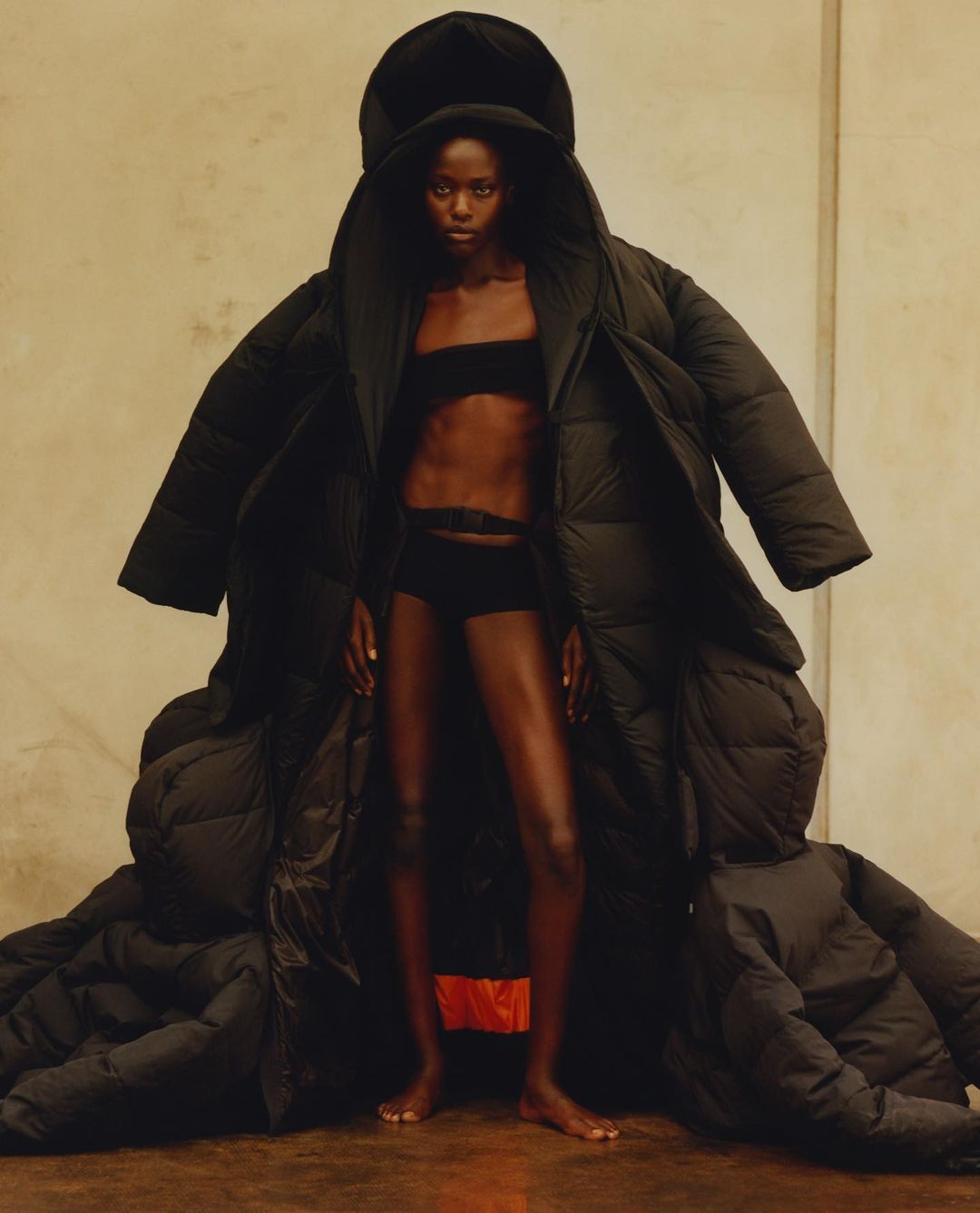 Fashion shot of Black woman in oversized black puffer coat, tube top, and briefs in studio with neutral background.