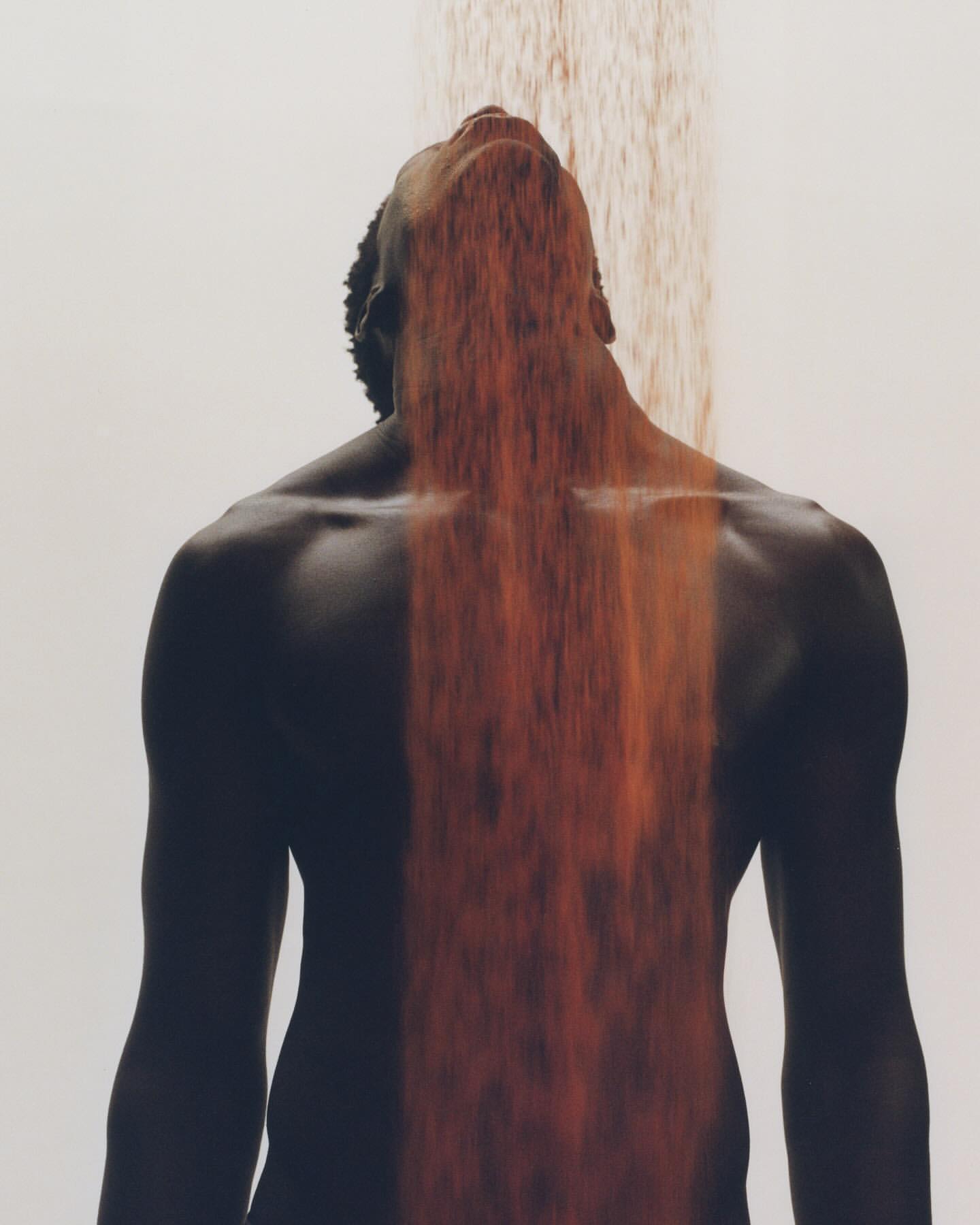 Artistic photograph of a dark-skinned shirtless adult male with face tilted upwards while being 'showered' with falling orange material.