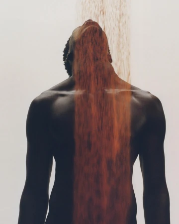 Artistic photograph of a dark-skinned shirtless adult male with face tilted upwards while being 'showered' with falling orange material.
