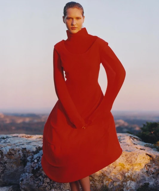 Elegant woman in red dress poses on rocky landscape against a pale sky. Fashion shot with natural light, minimalist style.