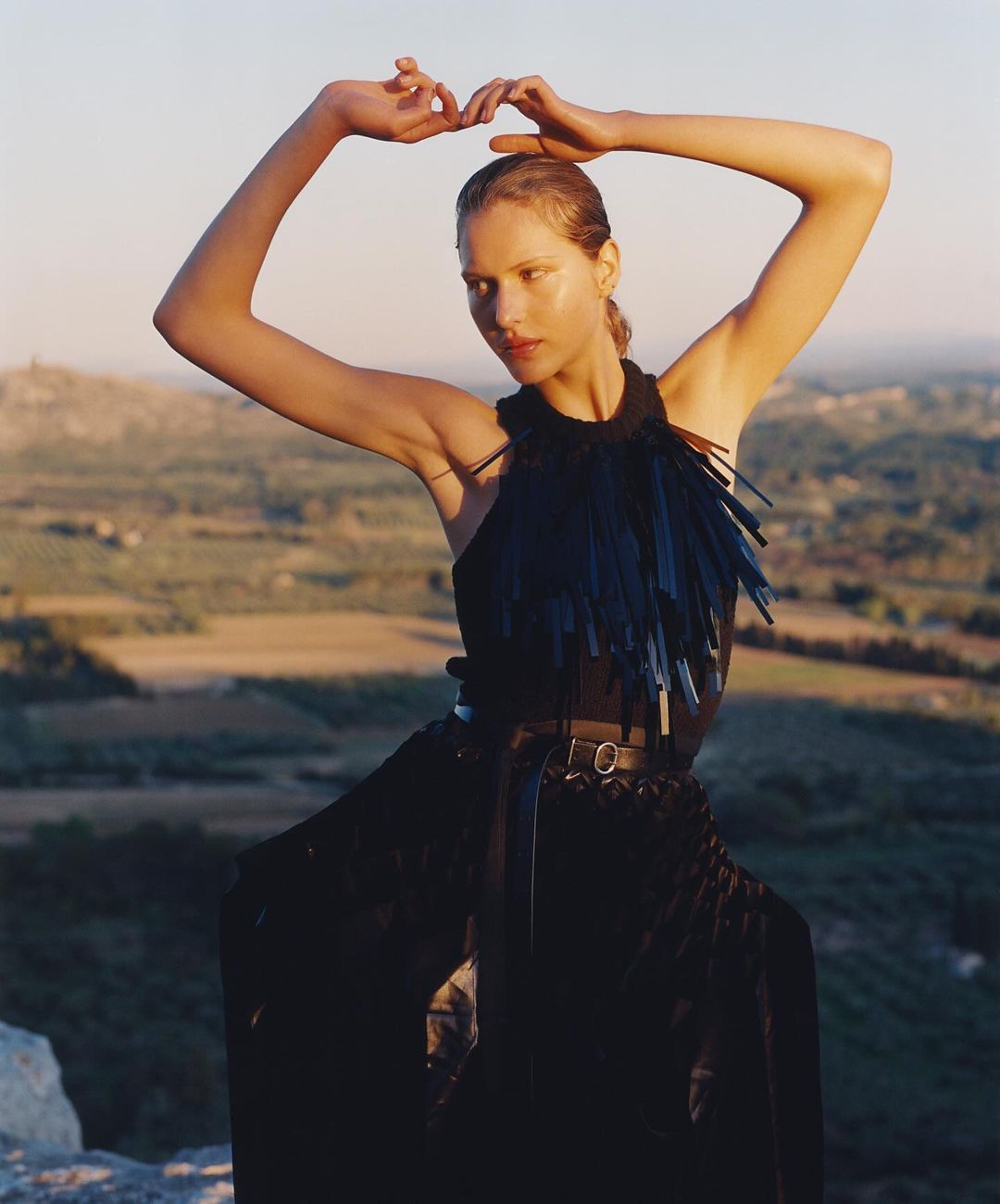 Adult woman poses in black dress in a sunlit, rural setting with distant hills. Fashion editorial portrait features an artistic composition.