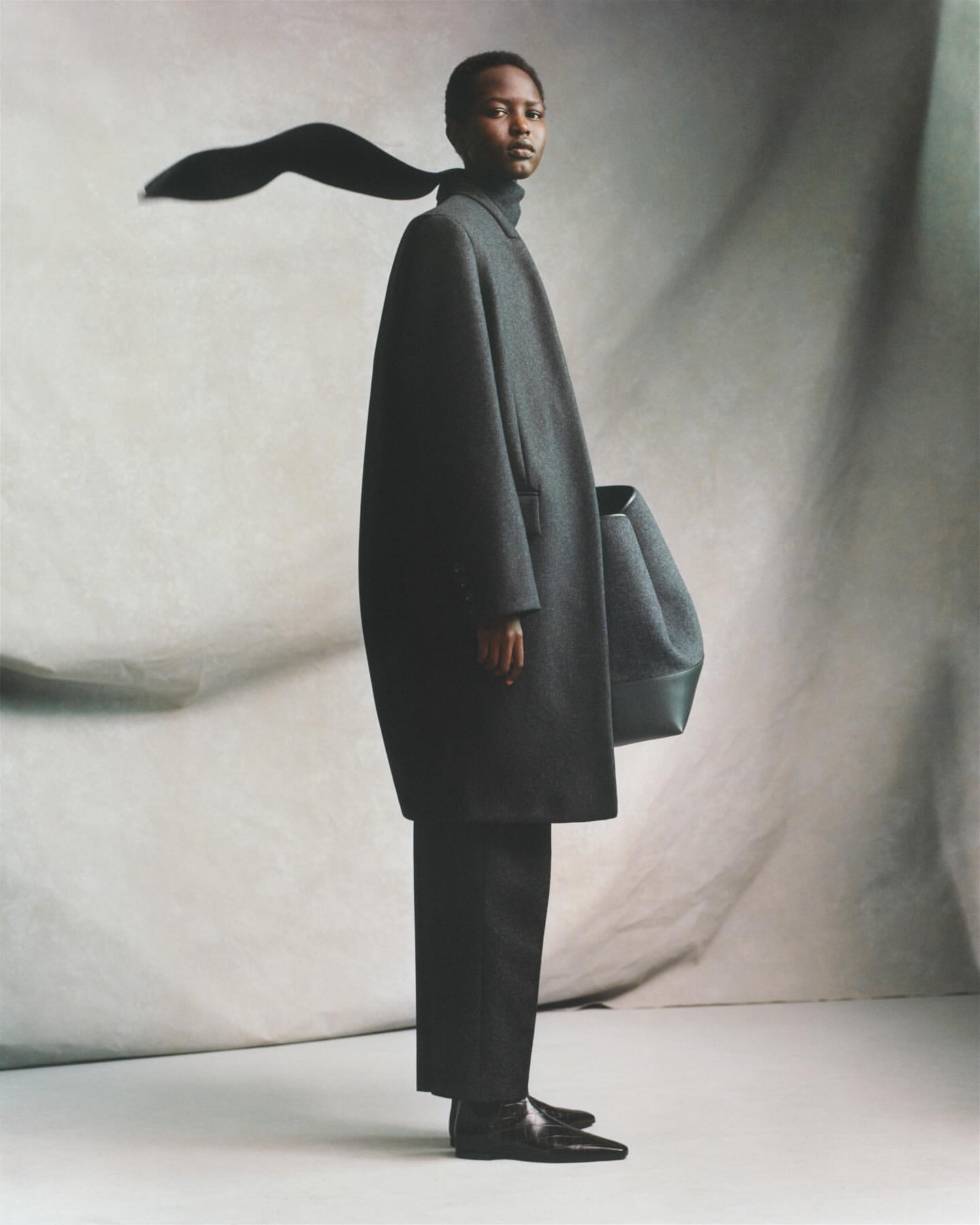 Fashion shot of a black female model in an oversized grey coat and black pants posing against a mottled grey backdrop in a studio.