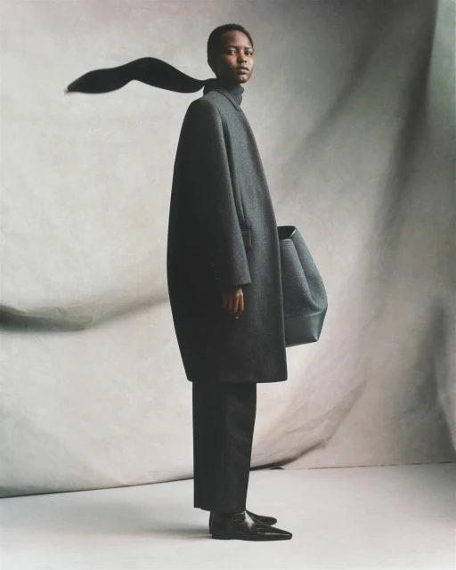 Fashion shot of a black female model in an oversized grey coat and black pants posing against a mottled grey backdrop in a studio.