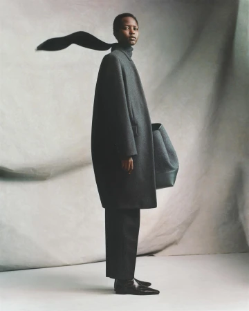 Fashion shot of a black female model in an oversized grey coat and black pants posing against a mottled grey backdrop in a studio.