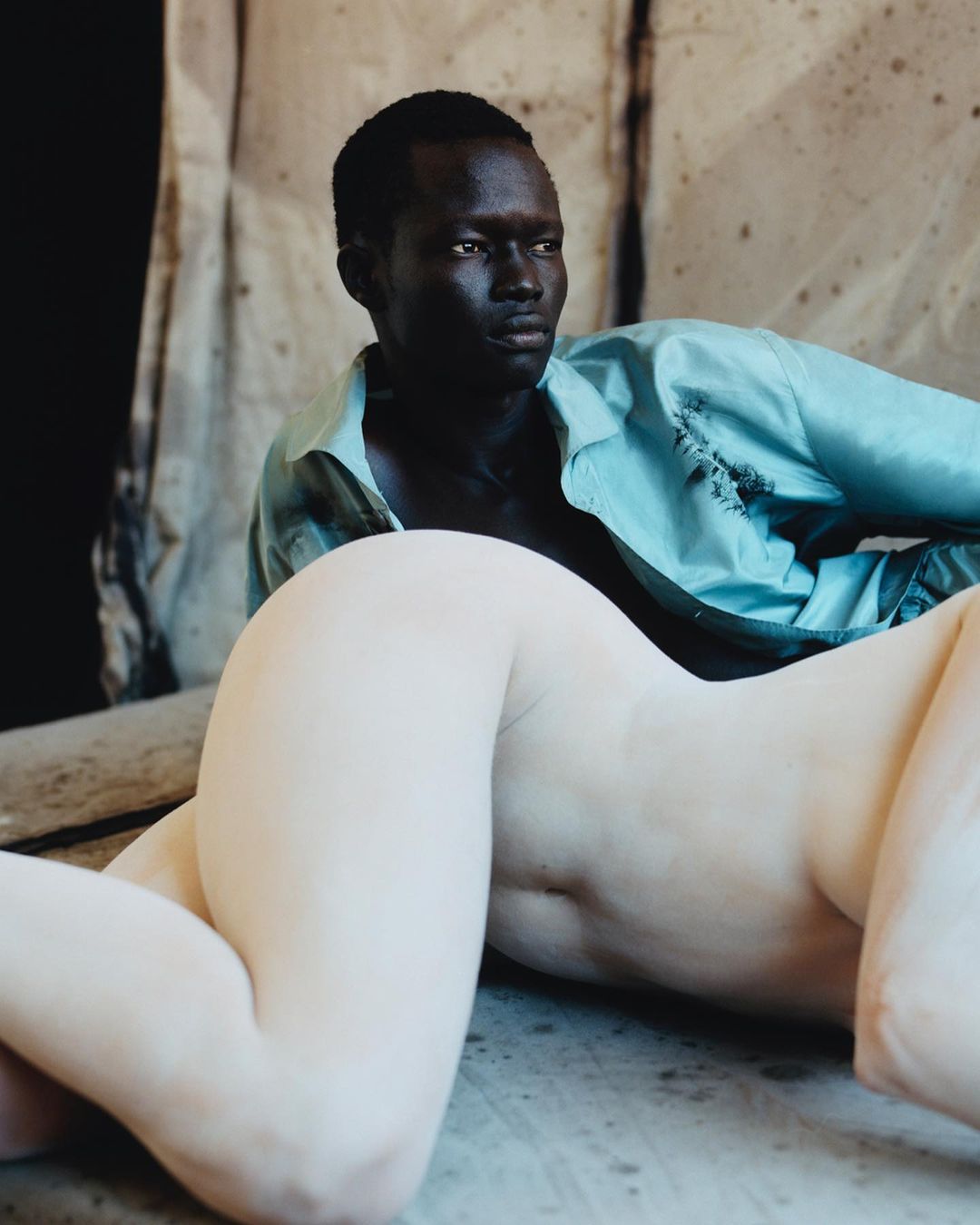 Man and mannequin in artistic studio portrait. Dark-skinned male with short hair poses near reclining pale female mannequin.