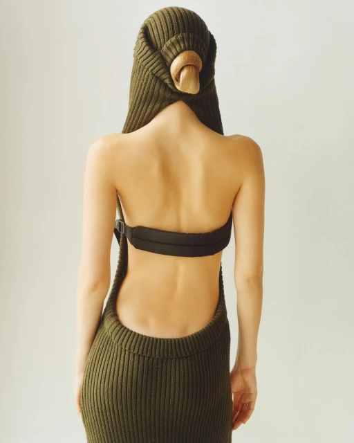 Back view of a woman wearing an olive green knit dress with a hood, a black bandeau top, and a wooden clasp, against a white background.
