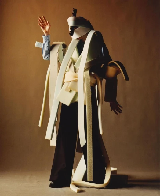 Person in elaborate foam costume poses in studio. Abstract fashion with headpiece, jacket, wide pants, and textured layers. 