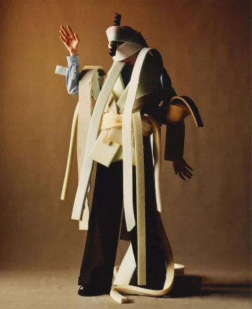 Person in elaborate foam costume poses in studio. Abstract fashion with headpiece, jacket, wide pants, and textured layers. 