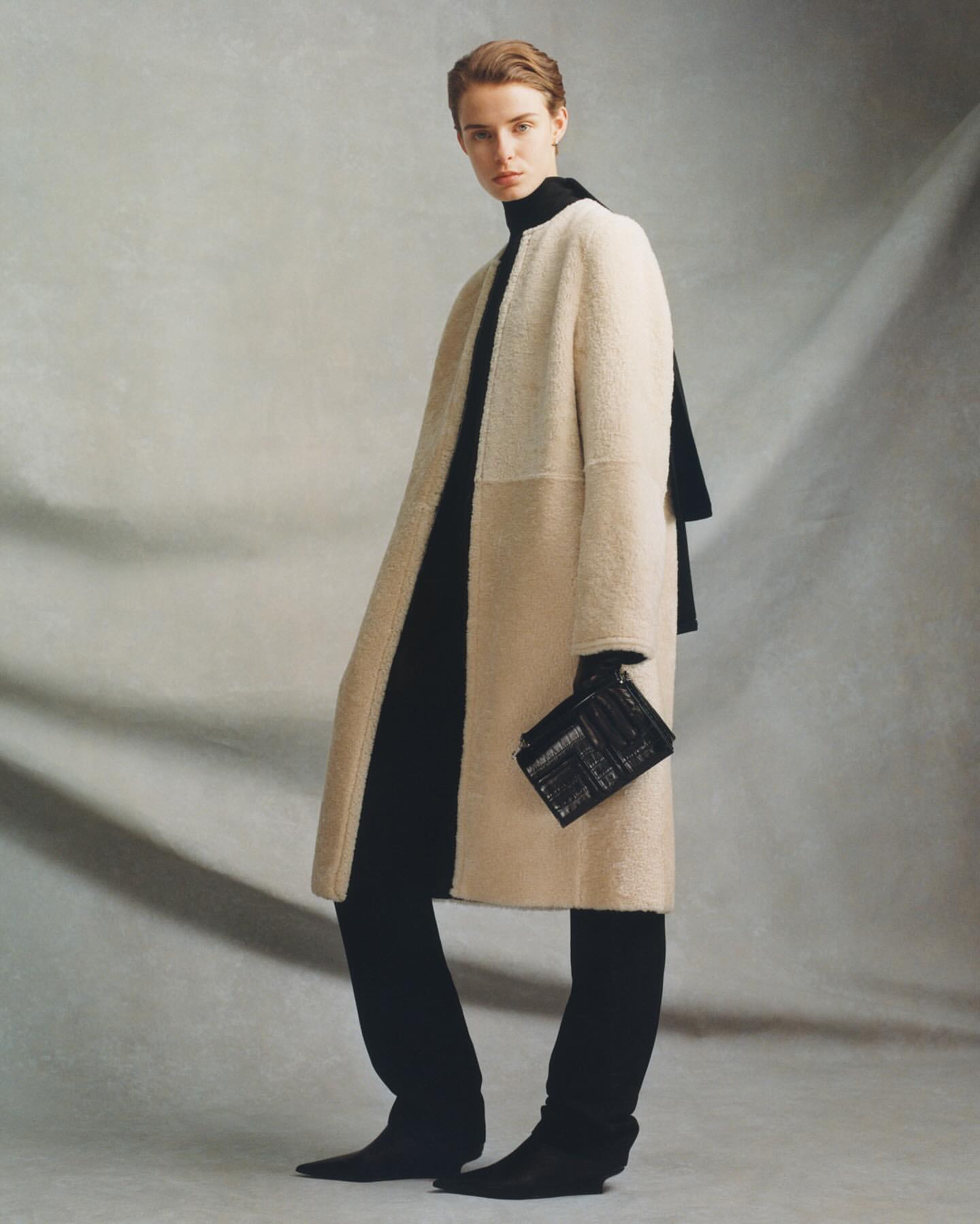 Model in a cream shearling coat wearing black trousers and holding a small black clutch against a textured grey backdrop.