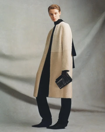 Model in a cream shearling coat wearing black trousers and holding a small black clutch against a textured grey backdrop.