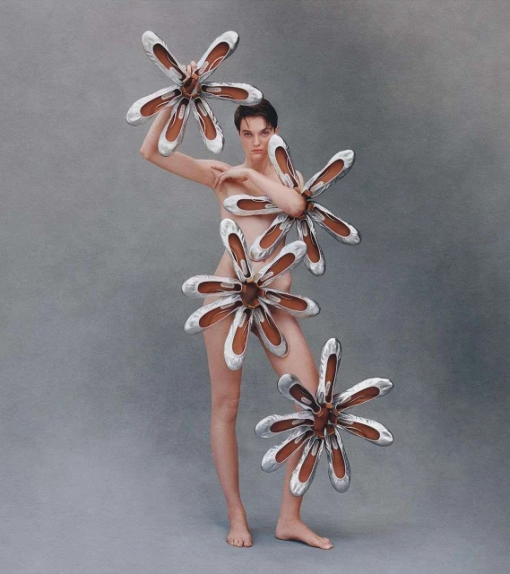 Artistic nude woman posing with flower-shaped ballet shoe arrangements in a photo studio against a neutral grey backdrop.