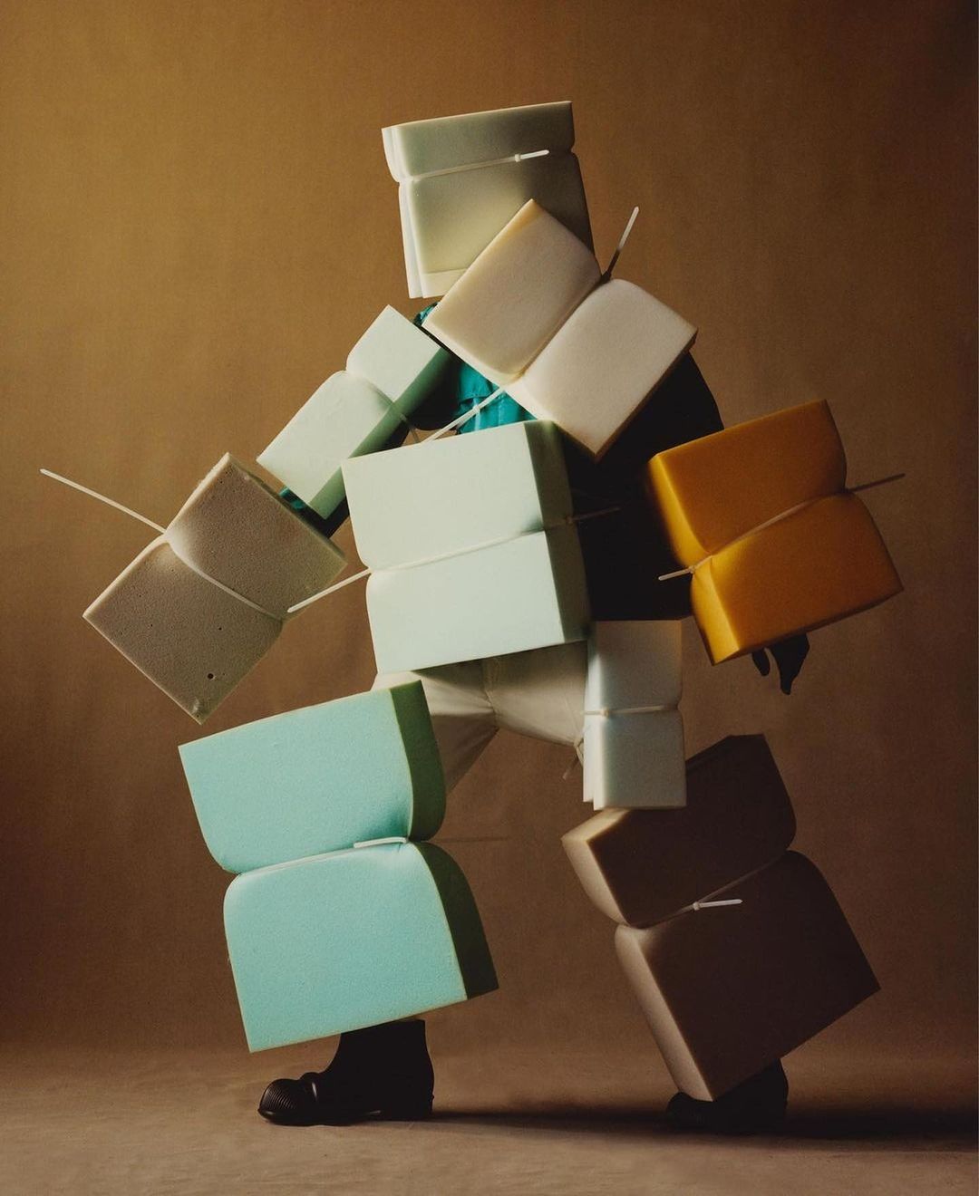 Person in a full body suit made of foam blocks in white, yellow, and turquoise in a studio with tan background. Conceptual fashion.