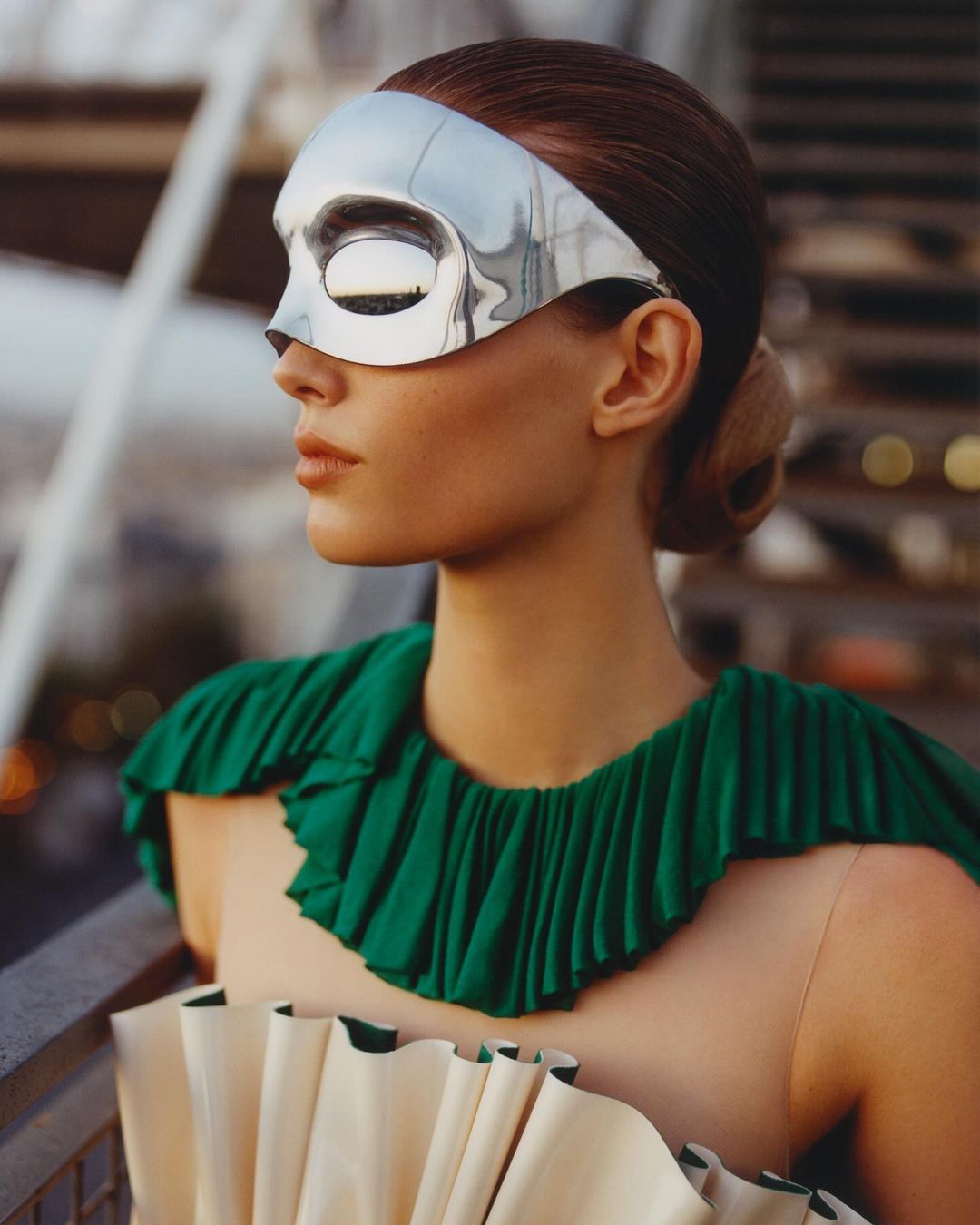 Woman in green ruffled collar and silver mask stands outdoors with city lights blurred in the distance, eye-level angle, medium shot.