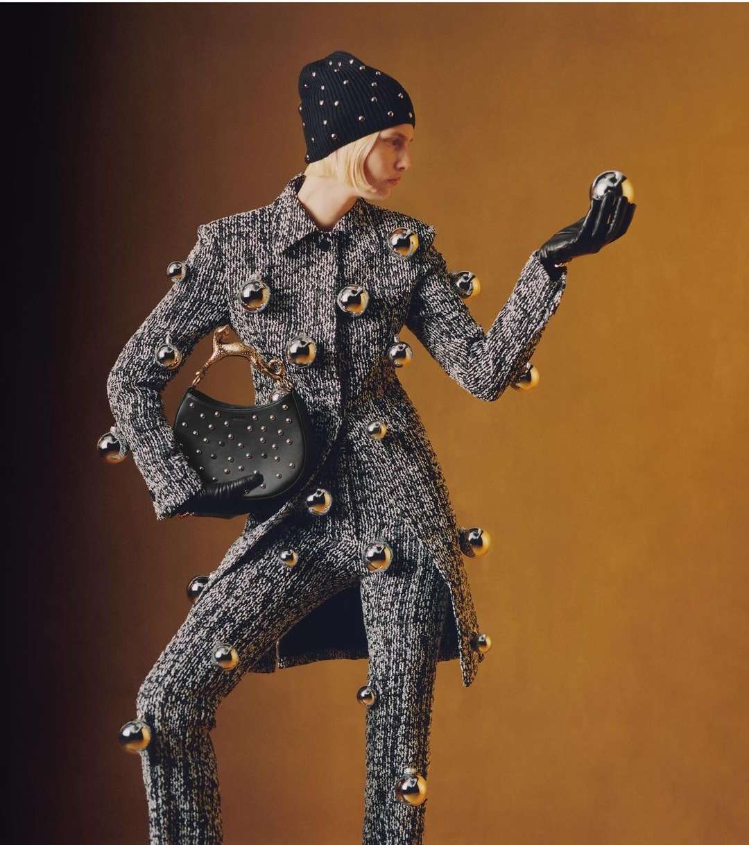Fashion photo of a model in a tweed suit adorned with silver spheres, posing with a purse and wearing a matching beanie and gloves.