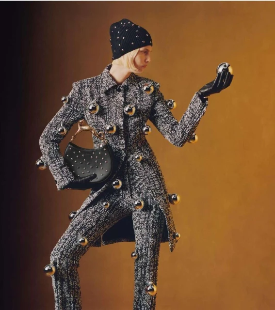 Fashion photo of a model in a tweed suit adorned with silver spheres, posing with a purse and wearing a matching beanie and gloves.
