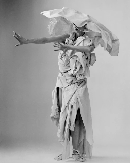 Monochrome fashion portrait of a person wearing an ensemble made of paper, posing with outstretched arms in a studio setting.