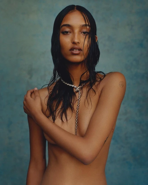 Striking studio portrait of a bare-chested woman with wet hair, posing confidently against a textured blue backdrop with a delicate chain necklace.