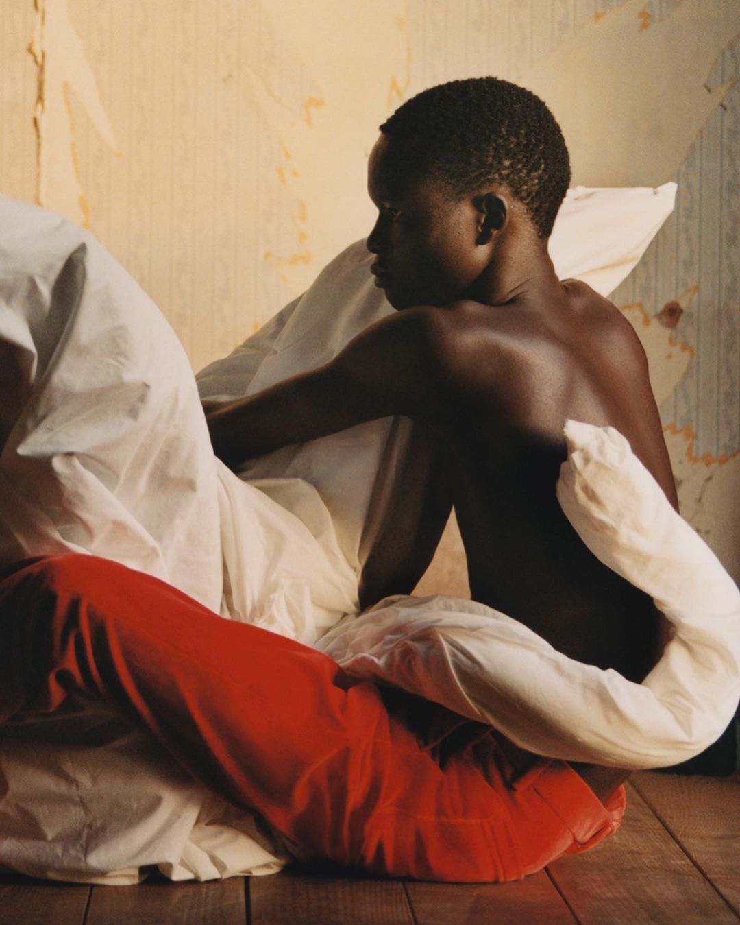 Young African boy sits indoors among white sheets and a red fabric, creating a portrait-style composition with warm, natural light.