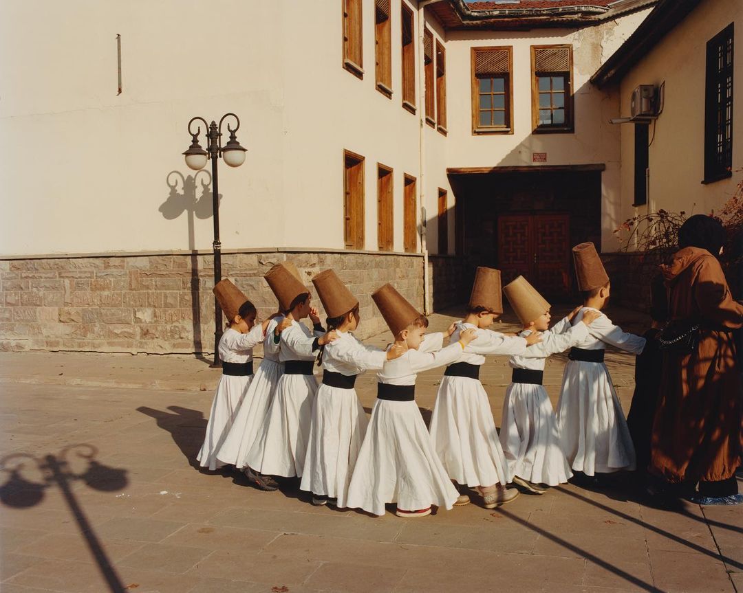 Children in traditional attire participate in a whirling dervish ceremony outdoors on a paved surface, with a building in the background.