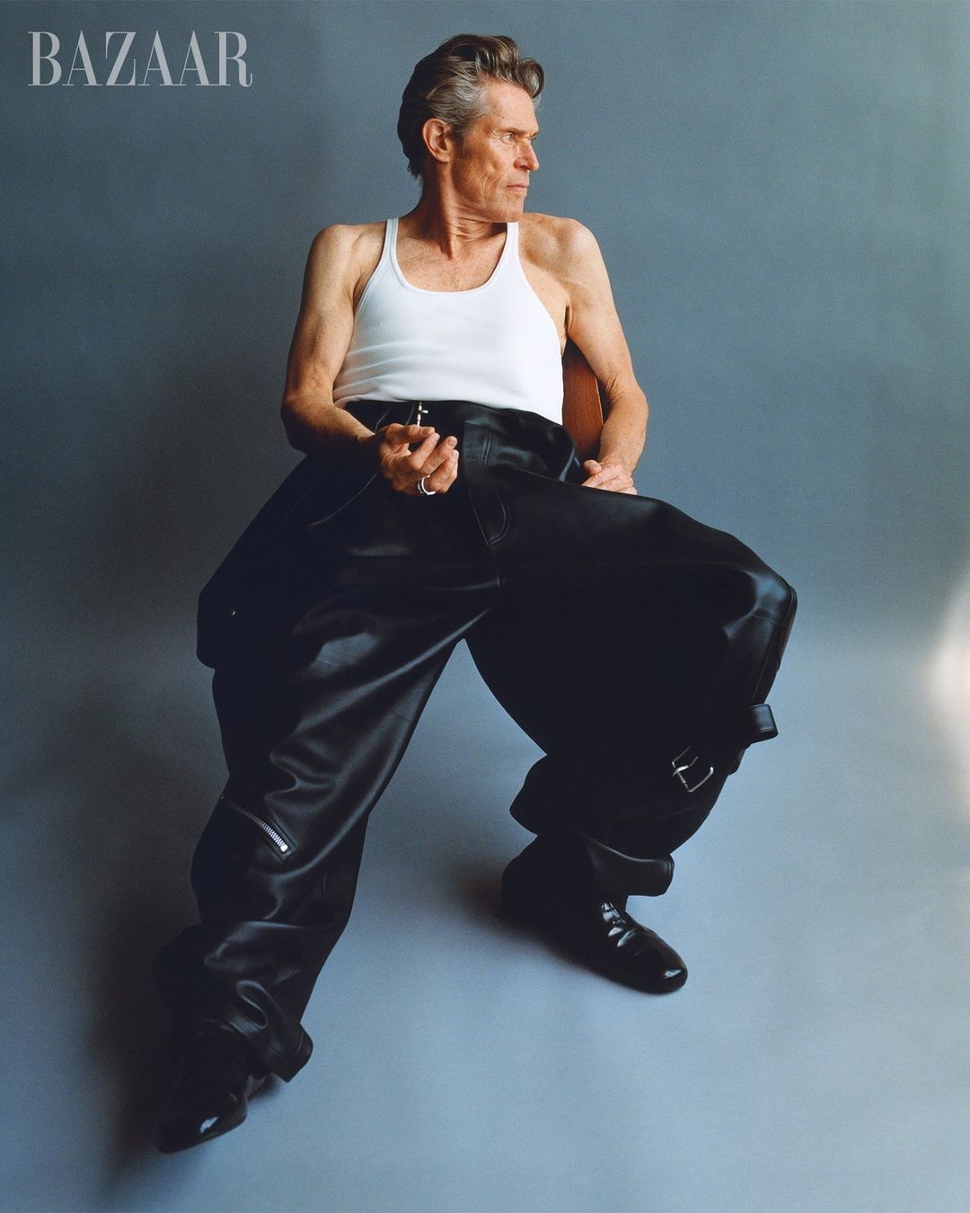 Willem Dafoe sits in a photo studio wearing a white tank top and oversized black leather pants and boots for Bazaar magazine.
