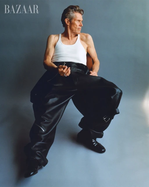 Willem Dafoe sits in a photo studio wearing a white tank top and oversized black leather pants and boots for Bazaar magazine.