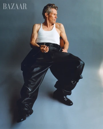 Willem Dafoe sits in a photo studio wearing a white tank top and oversized black leather pants and boots for Bazaar magazine.
