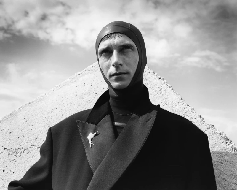 Portrait of an adult male wearing a swim cap, jacket, and brooch in front of a sky and a geometric concrete structure.