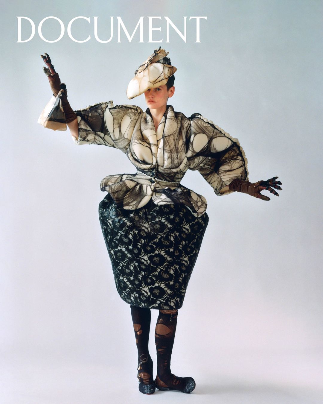 Fashion shot: a person in a sculptural outfit with a black floral skirt, a matching hat, and long gloves, posing on a light background.