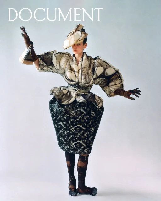 Fashion shot: a person in a sculptural outfit with a black floral skirt, a matching hat, and long gloves, posing on a light background.