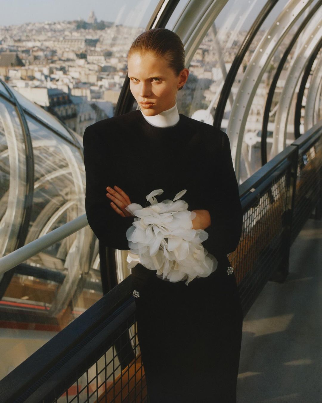 Elegant woman in black dress posing with arms crossed on a rooftop overlooking a city skyline, featuring modern architecture and neutral tones.