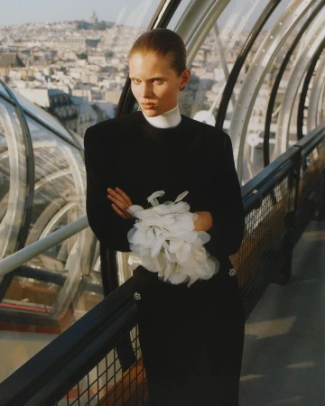 Elegant woman in black dress posing with arms crossed on a rooftop overlooking a city skyline, featuring modern architecture and neutral tones.