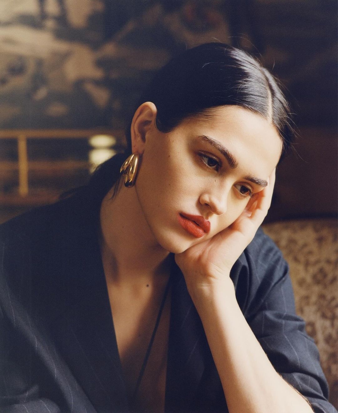 Portrait of an adult female with red lipstick and hoop earrings wearing a pinstripe jacket, resting her head on her hand indoors.