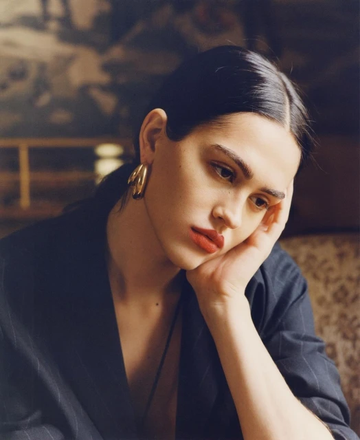 Portrait of an adult female with red lipstick and hoop earrings wearing a pinstripe jacket, resting her head on her hand indoors.