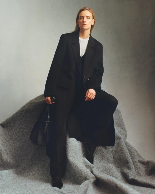 A fair-skinned woman in a black pantsuit and white top poses in a studio setting with a black purse. She sits on a gray cloth.