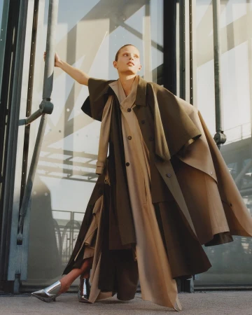 Fashionable woman poses in earth-toned layered coat in an urban outdoor setting near reflective building with metal framework.