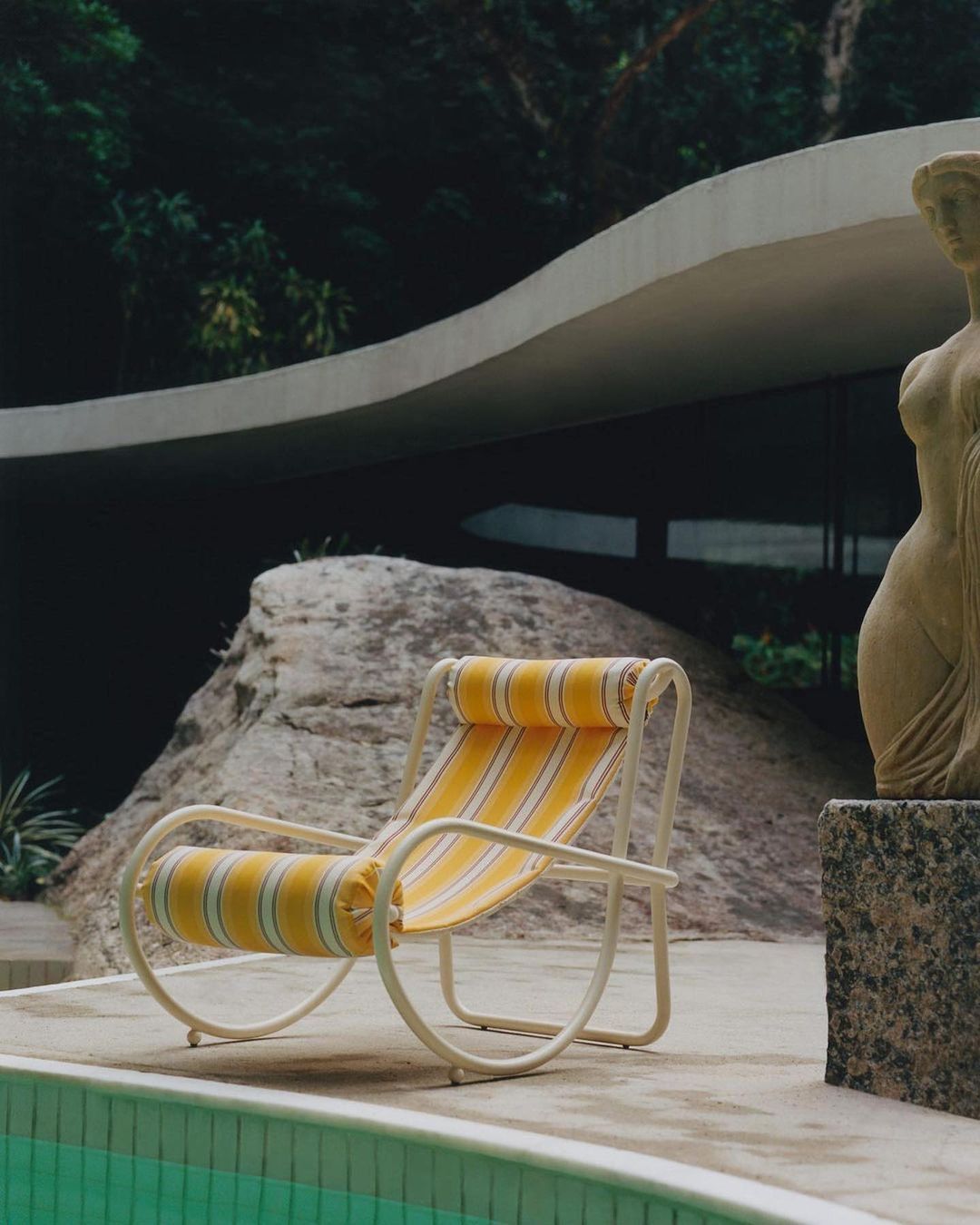Vintage-style outdoor scene featuring a striped yellow and white lounge chair next to a pool and a stone sculpture of a woman.