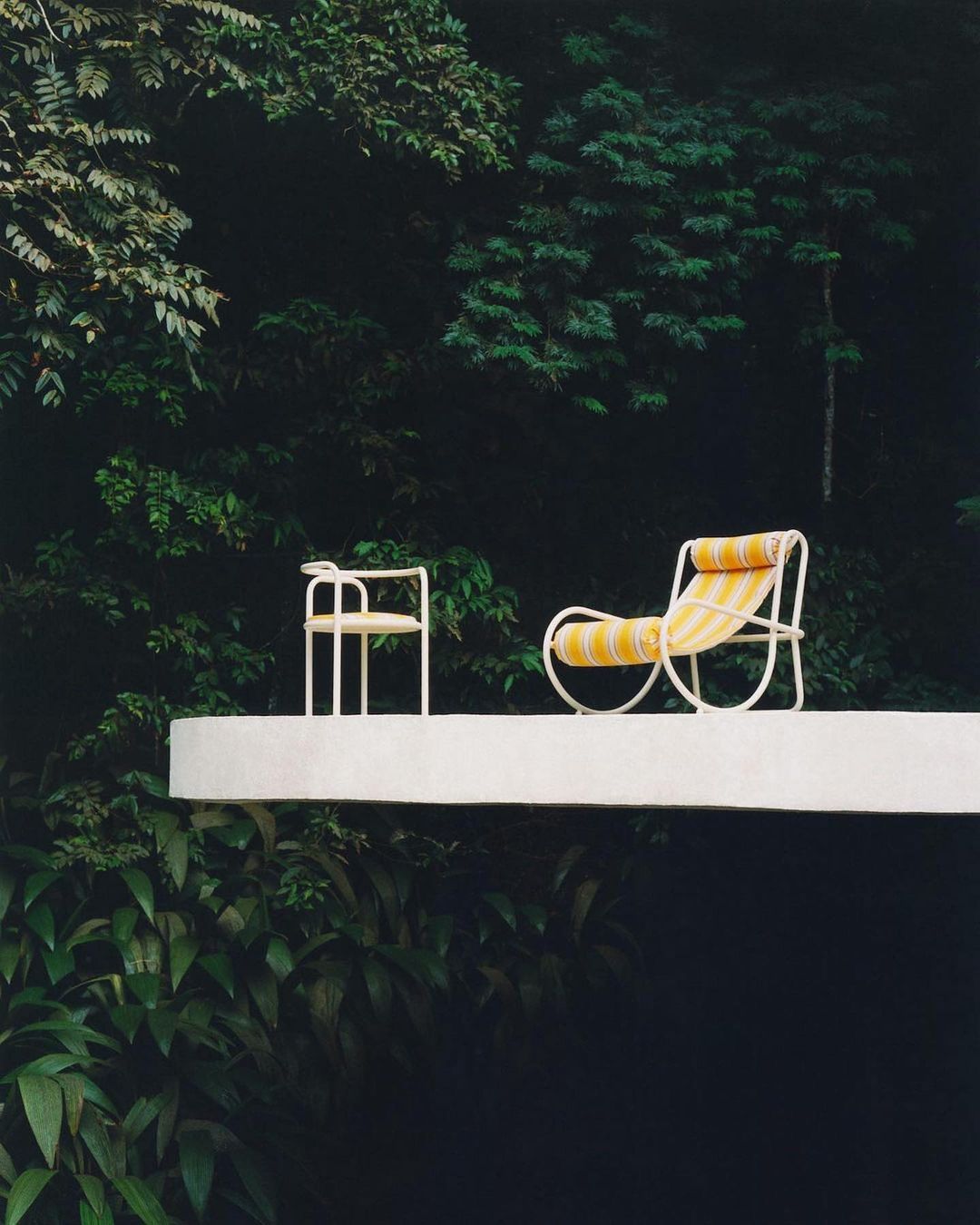 Outdoor furniture on a white patio with a chair and lounger, set against lush green foliage for a minimalist outdoor living space.